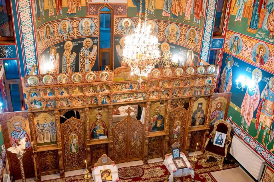 Interiorul unei biserici prezintă un iconostas din lemn sculptat decorat cu picturi religioase și un candelabru suspendat în centru.