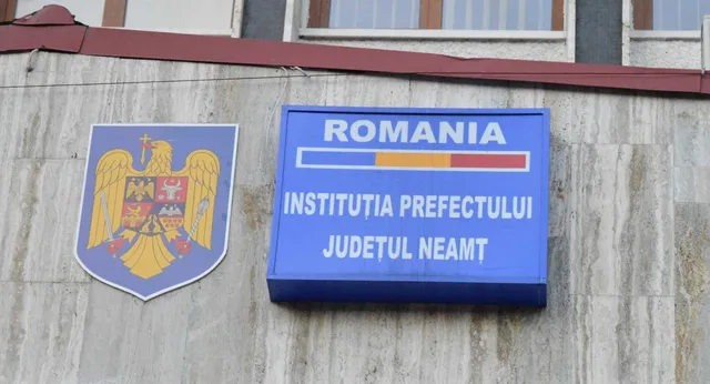 Un panou albastru cu textul ROMANIA INSTITUȚIA PREFECTULUI JUDEȚUL NEAMȚ și un scut cu un vultur sunt montate pe un perete gri.