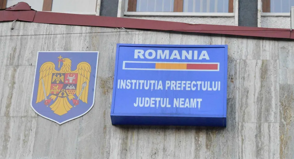 Un panou albastru cu textul ROMANIA INSTITUȚIA PREFECTULUI JUDEȚUL NEAMȚ și un scut cu un vultur sunt montate pe un perete gri.