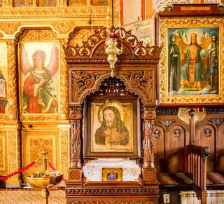 În interiorul unei biserici, o icoană cu o figură aurie este încadrată în lemn sculptat, lângă alte picturi religioase și un sfeșnic metalic.