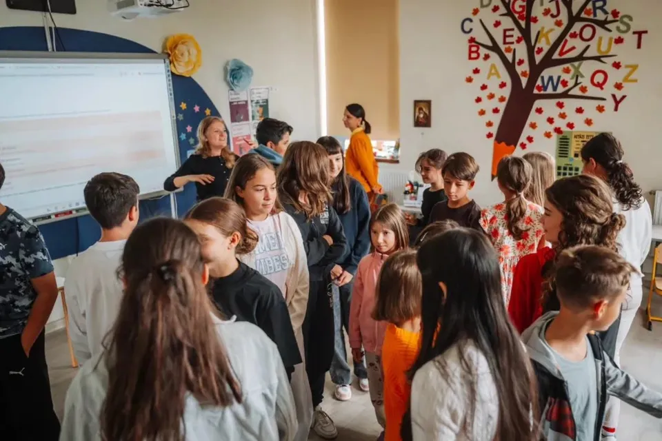 Un grup de copii stă în picioare într-o sală de clasă, orientat spre o femeie care stă lângă un ecran de proiecție alb.