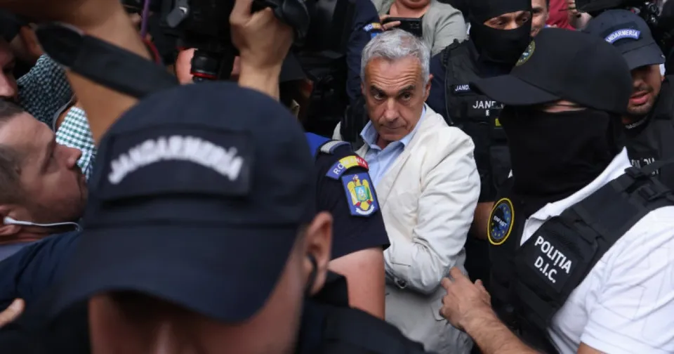 Un bărbat cu părul cărunt și sacou deschis este înconjurat de persoane purtând uniforme și căști de poliție.