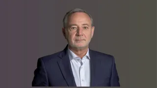 Un bărbat cu părul scurt și gri poartă un sacou închis la culoare peste o cămașă albă, pe un fundal gri uniform.