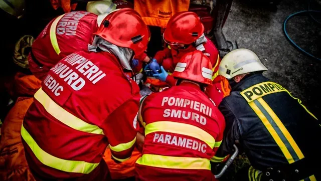 Patru persoane purtând echipamente de protecție de culoare roșie și una în echipament negru cu benzi galbene stau grupate într-un spațiu îngust.