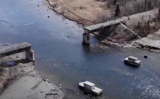 O vedere de sus prezintă un pod de beton parțial rupt peste un râu, cu piloni și secțiuni de structură vizibile în apă și pe maluri.