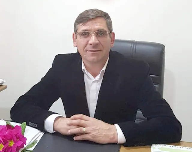 Un bărbat cu ochelari și sacou negru stă la un birou cu mâinile încrucișate, purtând un inel la degetul inelar.