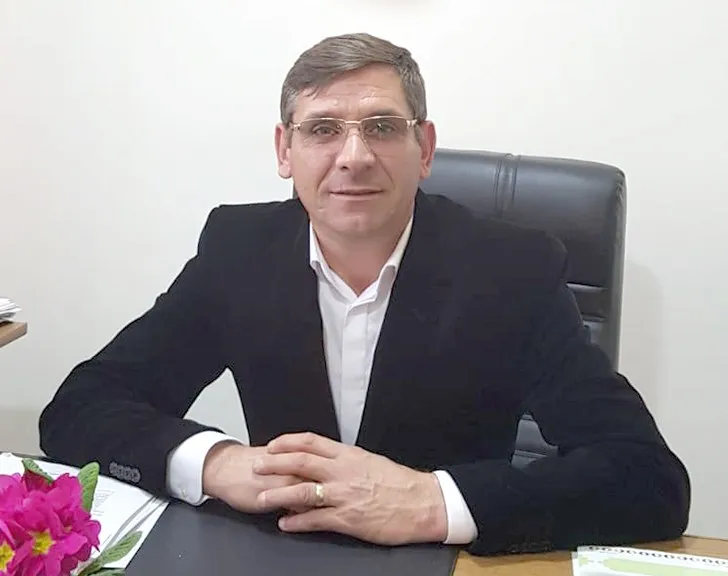 Un bărbat cu ochelari și sacou negru stă la un birou cu mâinile încrucișate, purtând un inel la degetul inelar.