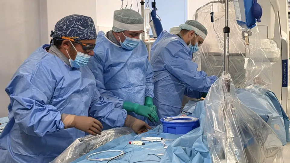 Trei persoane în halate medicale albastre și măști de protecție lucrează lângă un pacient acoperit în cadrul unei intervenții chirurgicale.