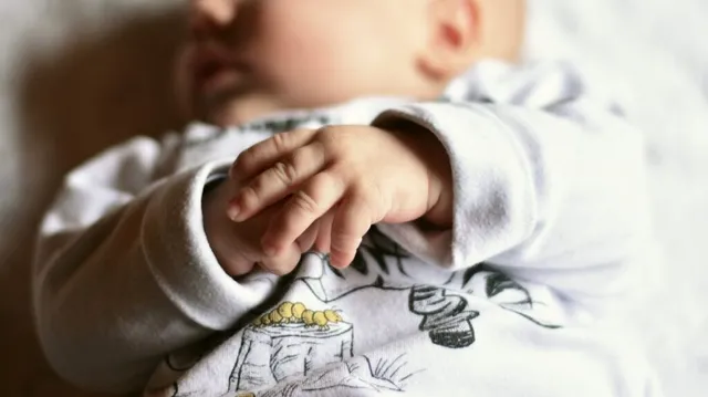 Un nou-născut purtând haine albe cu imprimeuri este întins pe o suprafață deschisă la culoare, având mâinile lipite una de alta în prim-plan.