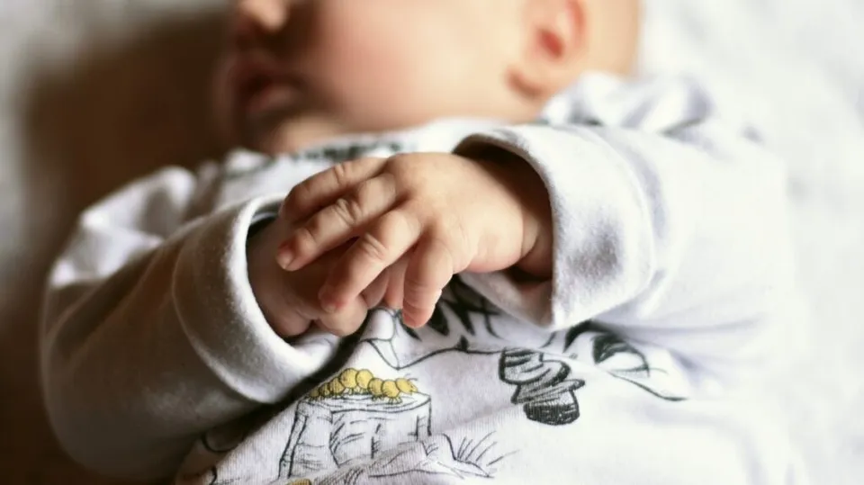 Un nou-născut purtând haine albe cu imprimeuri este întins pe o suprafață deschisă la culoare, având mâinile lipite una de alta în prim-plan.