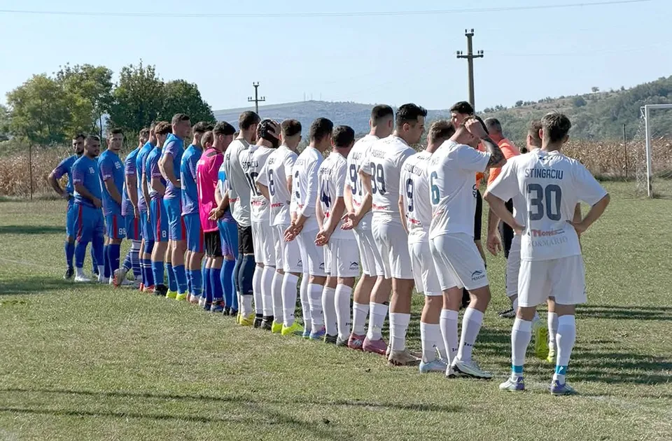 O linie de jucători de fotbal în echipamente albastre și albe stă întinsă pe un teren de iarbă sub un cer senin.