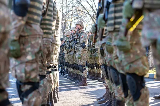 Soldați în uniforme de camuflaj și echipament de protecție stau în rând pe o stradă asfaltată sub lumina soarelui.