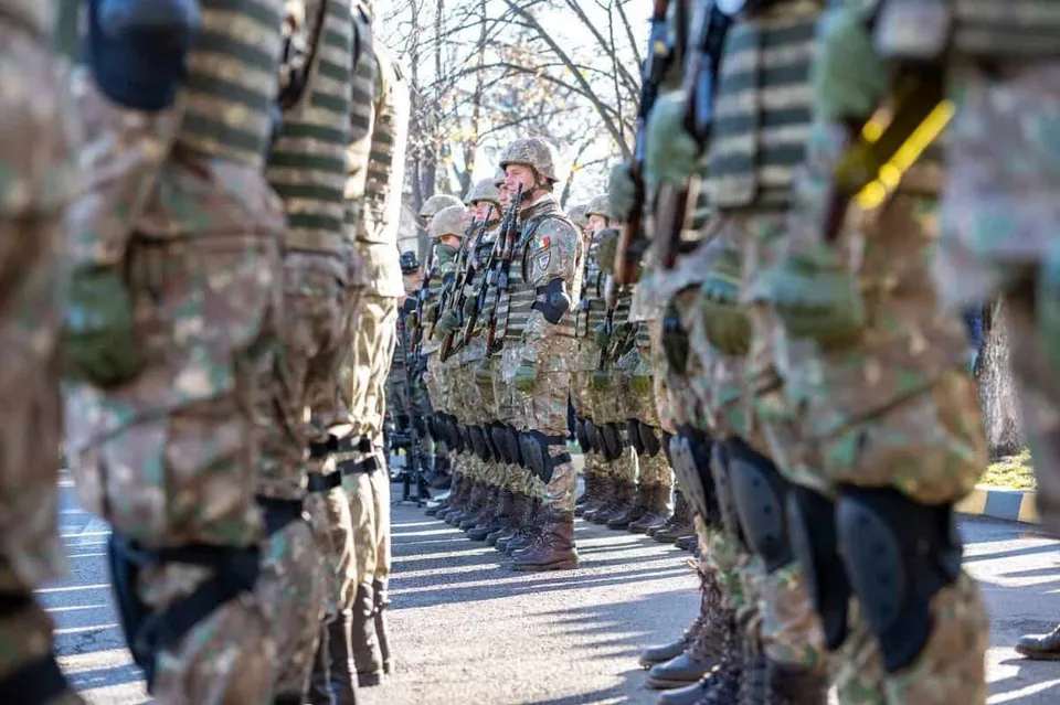 Soldați în uniforme de camuflaj și echipament de protecție stau în rând pe o stradă asfaltată sub lumina soarelui.