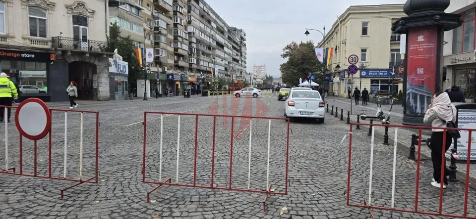 O stradă pavată cu pietriș este delimitată de bariere metalice roșii și albe, având mașini și oameni în fundal sub un cer înnorat.