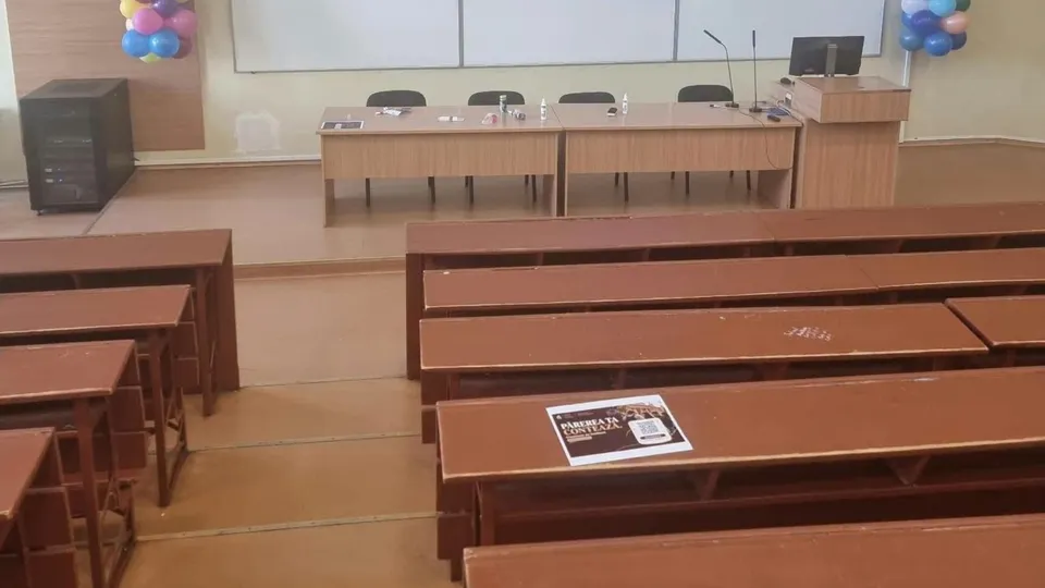 O sală de curs cu bănci de lemn maro, un birou lung în față și un monitor de calculator pe un suport de lemn.