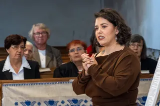 O femeie cu părul scurt și închis la culoare poartă o bluză maro și vorbește în timp ce un grup de persoane stă în fundal.