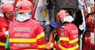 Trei pompieri în uniforme roșii cu benzi galbene transportă o persoană pe o targă în spatele unei ambulanțe.