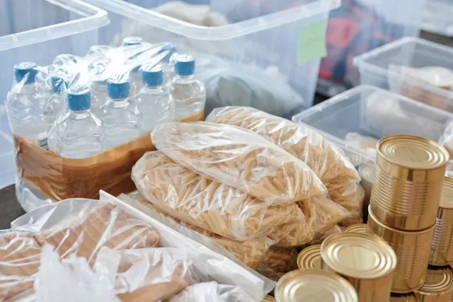 Mai multe pungi de plastic cu paste și cutii metalice de conserve sunt așezate pe suprafețe albe lângă sticle de apă transparente.