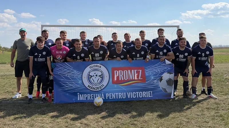 Un grup de bărbați în echipamente sportive de fotbal stau în rând în fața unui banner albastru pe un teren de fotbal sub cerul senin.
