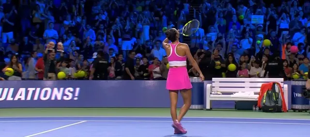 O femeie cu o rochie roz și un tricou roz privește spre tribunele unui teren de tenis.