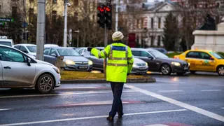 Un polițist cu vestă reflectorizantă verde reglează traficul pe un drum urban cu mai multe mașini în fundal.