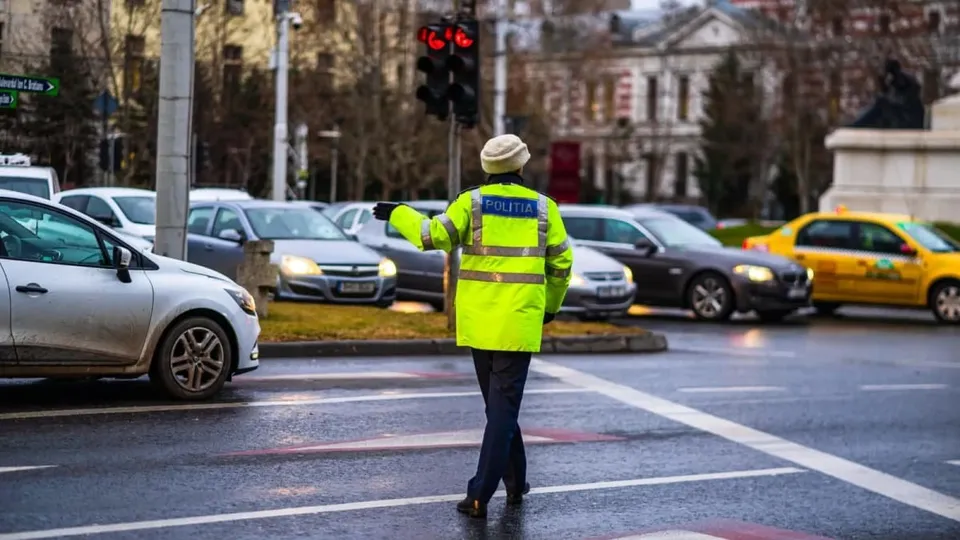 Un polițist cu vestă reflectorizantă verde reglează traficul pe un drum urban cu mai multe mașini în fundal.