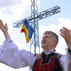 Un bărbat cu mâinile ridicate poartă o cămașă albă brodată și o vestă colorată sub un steag și o structură metalică înaltă.