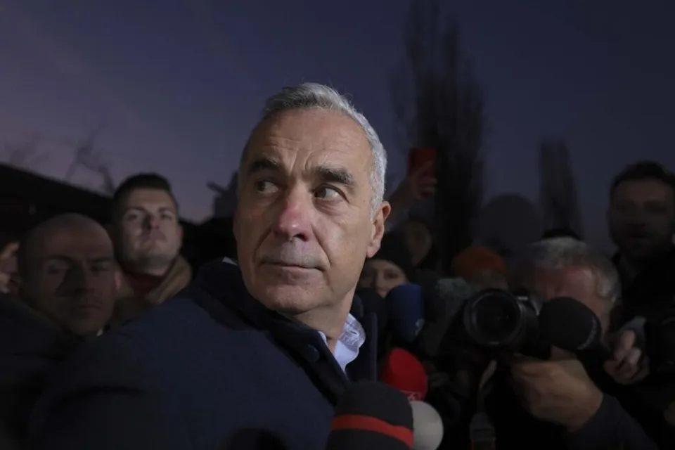 Un bărbat cu părul scurt și cămăși deschizând la guler este înconjurat de microfoane și persoane în fundal întunecat.