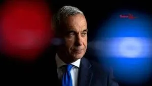 Un bărbat cu părul cărunt, purtând sacou închis și cravată albastră, este surprins în prim-plan cu lumini roșii și albastre difuze în fundal.