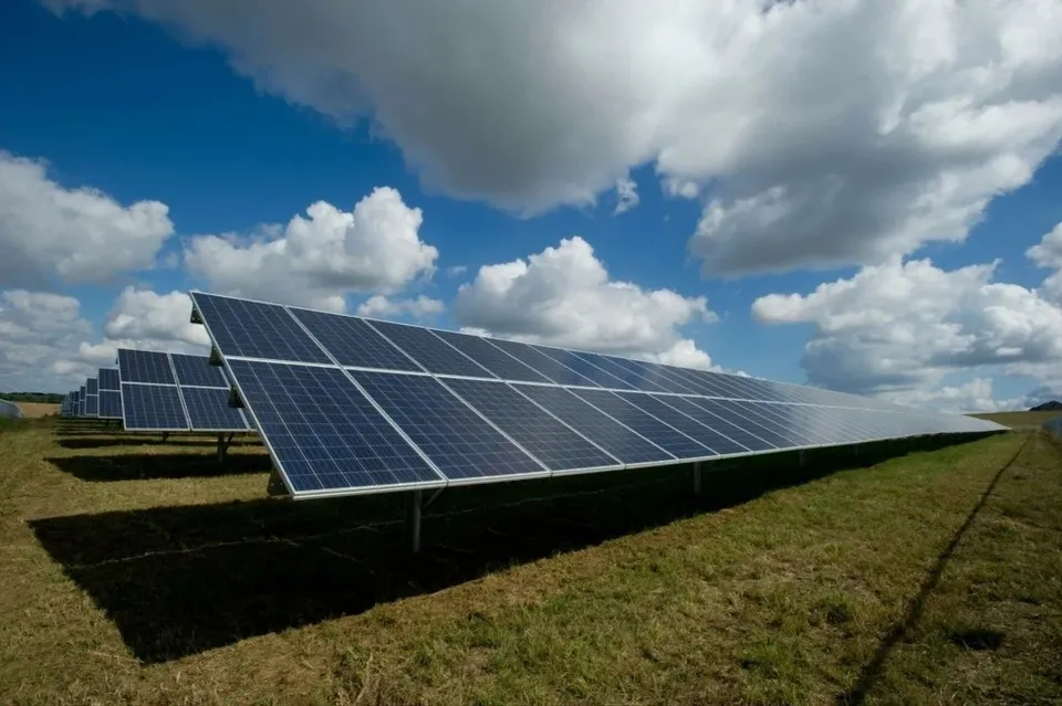 Rânduri de panouri fotovoltaice sunt instalate pe un câmp de iarbă sub un cer cu nori albi și albastru.