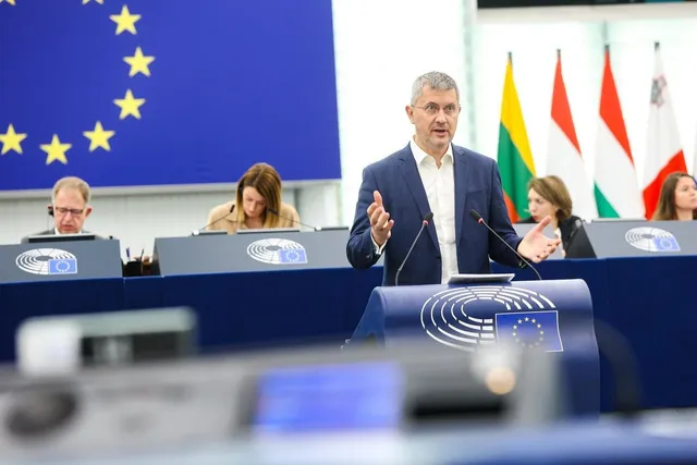 Un bărbat într-un sacou albastru stă în spatele unui pupitru cu un logo alb, gesticulând cu mâinile în fața unor microfoane.