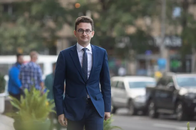Un bărbat cu ochelari și costum albastru stă în picioare pe un drum cu mașini și vegetație în fundal.