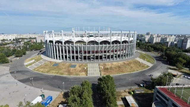 O vedere aeriană prezintă un stadion circular mare cu coloane verticale albe, înconjurat de un drum asfaltat și zone cu iarbă uscată.