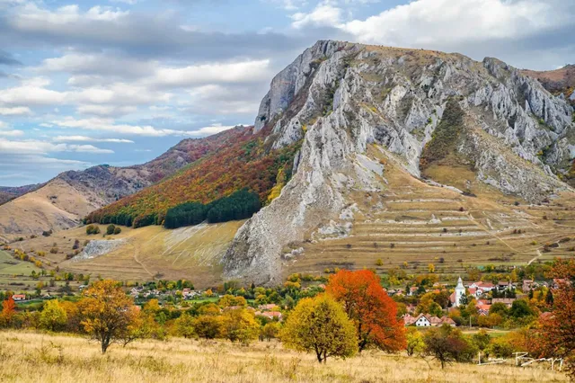 Un masiv muntos stâncos se ridică deasupra unui sat cu case cu acoperișuri roșii și vegetație cu frunziș colorat de toamnă.