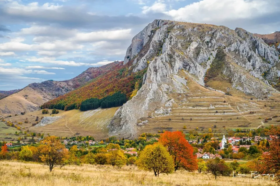Un masiv muntos stâncos se ridică deasupra unui sat cu case cu acoperișuri roșii și vegetație cu frunziș colorat de toamnă.
