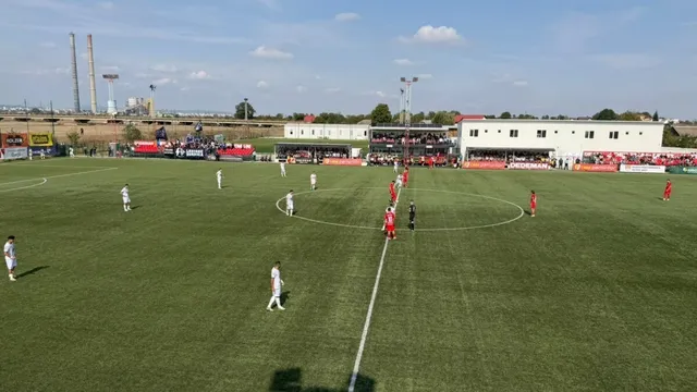 Jucători de fotbal în echipamente albe și roșii sunt dispersați pe un teren de fotbal verde sub un cer senin.