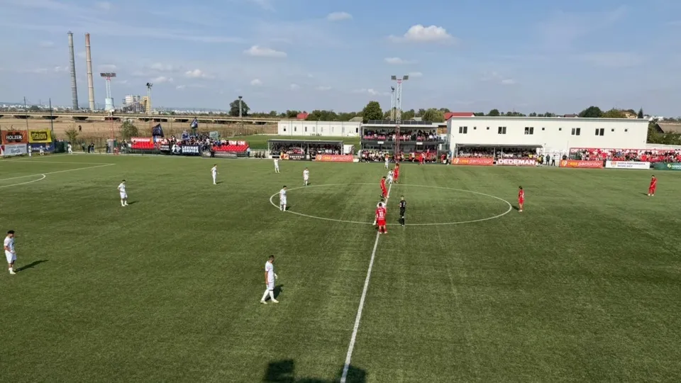 Jucători de fotbal în echipamente albe și roșii sunt dispersați pe un teren de fotbal verde sub un cer senin.