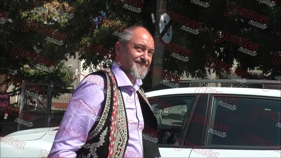 Un bărbat cu barbă și vestă decorată stă lângă o mașină albă în fața unor copaci.