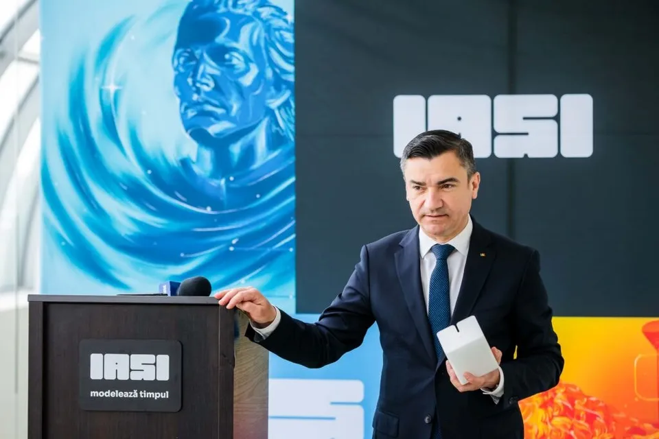 Un bărbat într-un costum închis și cravată albastră stă în fața unui podium negru, ținând un obiect alb în mână.