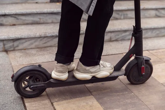 O persoană cu pantaloni negri și încălțăminte gri stă pe o trotinetă electrică neagră aflată pe un trotuar de piatră lângă niște trepte.