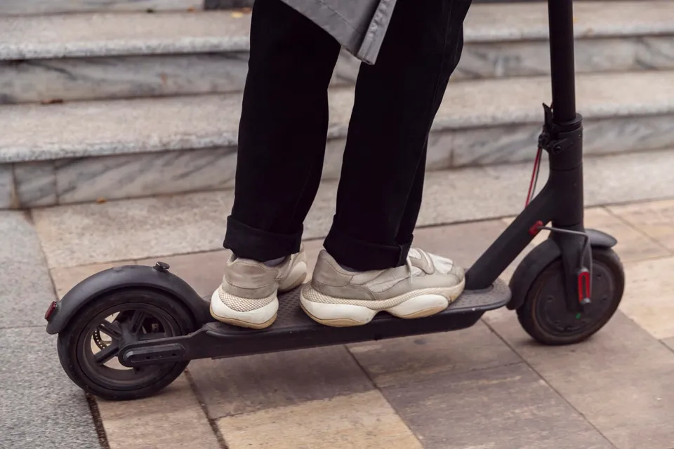 O persoană cu pantaloni negri și încălțăminte gri stă pe o trotinetă electrică neagră aflată pe un trotuar de piatră lângă niște trepte.