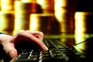 O mână apasă pe tastele unui tastatură negru de lângă un ecran de laptop, în timp ce stive de monede aurii sunt în fundal.
