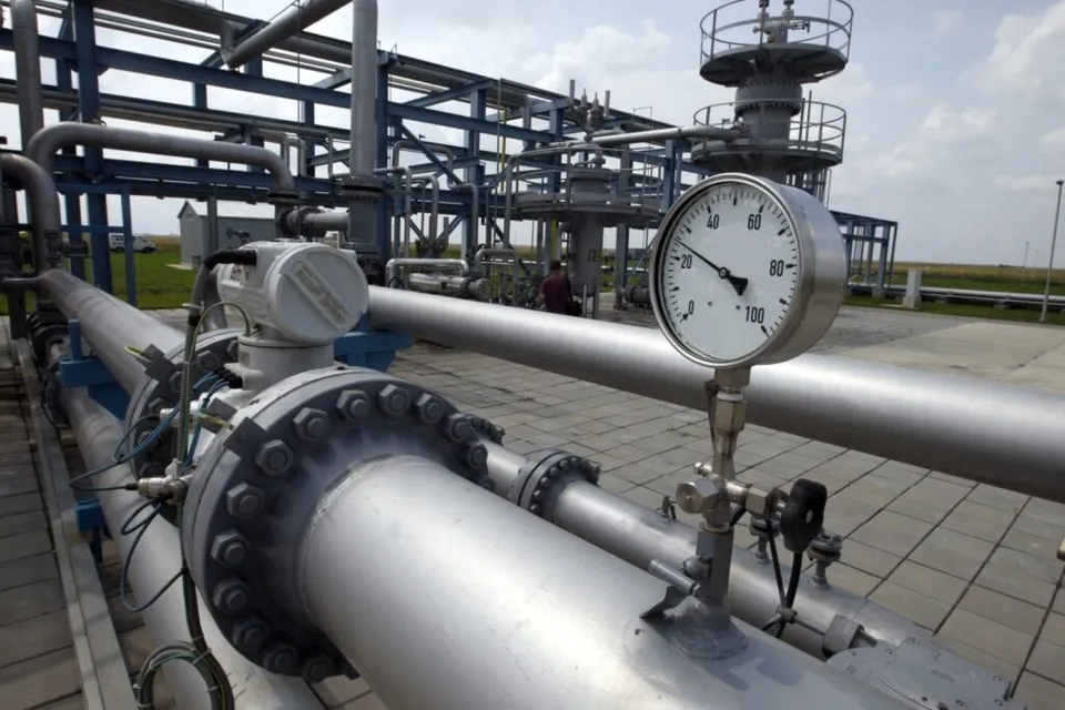 Conducte metalice gri și un manometru circular sunt instalate într-o stație industrială sub un cer înnorat.