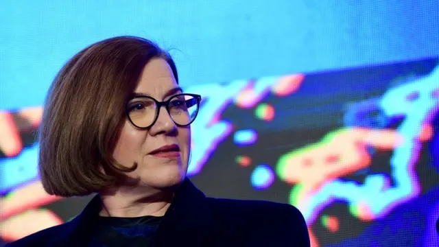 O femeie cu ochelari și păr scurt, brunet, poartă o vestă neagră în fața unui fundal colorat și luminat.