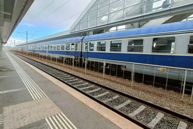 Un tren albastru și gri stă pe șine lângă un peron de beton cu linii de siguranță albe, sub o structură modernă de sticlă și metal.