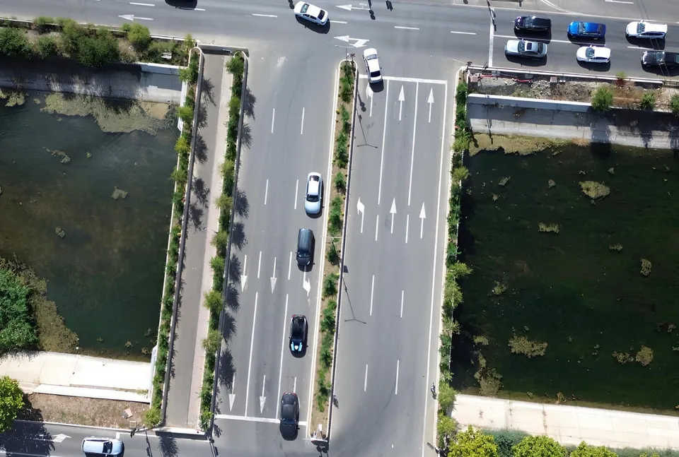 O vedere aeriană prezintă o porțiune de drum cu mașini circulând și parcate, flancată de două corpuri de apă și vegetație.