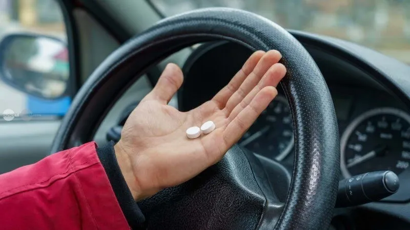 O mână ține două capsule albe deasupra unui volan de mașină.