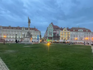O coloană înaltă cu sculpturi se ridică într-o piață cu un câmp de iarbă verde și clădiri colorate în fundal sub un cer înnorat.