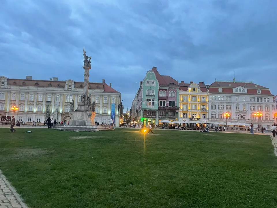O coloană înaltă cu sculpturi se ridică într-o piață cu un câmp de iarbă verde și clădiri colorate în fundal sub un cer înnorat.