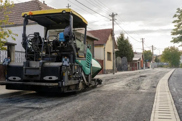 O mașină de asfaltat neagră și verde stă pe un drum de pietriș lângă un trotuar nou și case de proprietate.